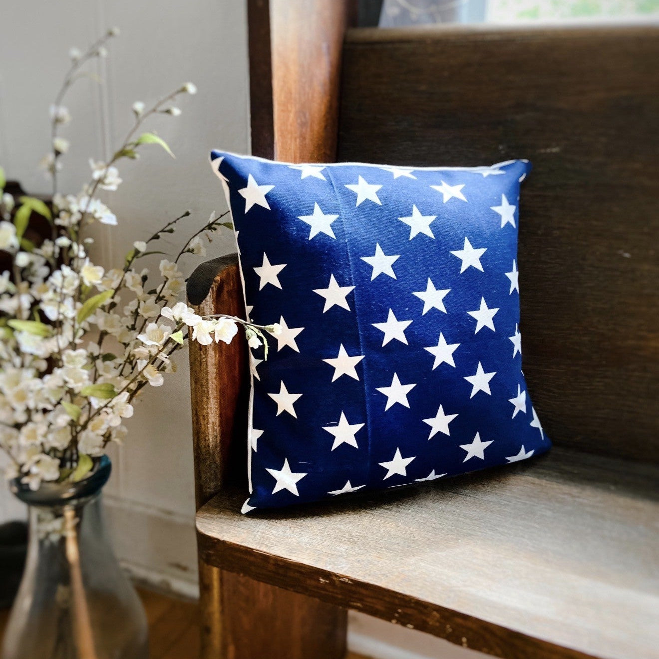 Blue pillow with white star pattern on a wooden bench next to a vase with flowers.