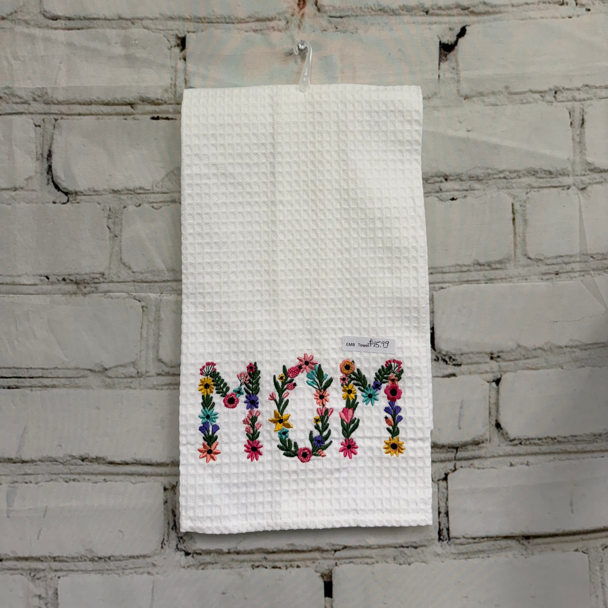 Embroidered towel with 'MOM' in colorful letters hanging on a brick wall.