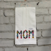 Embroidered towel with 'MOM' in colorful letters hanging on a brick wall.