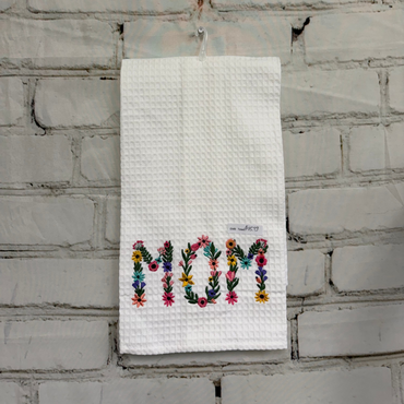 Embroidered towel with 'MOM' in colorful letters hanging on a brick wall.