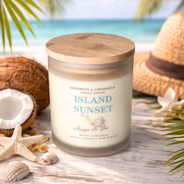 Candle labeled 'Island Sunset' from Coconuts & Cornbread Candle Company on a beach setting with coconuts, starfish, and a hat.